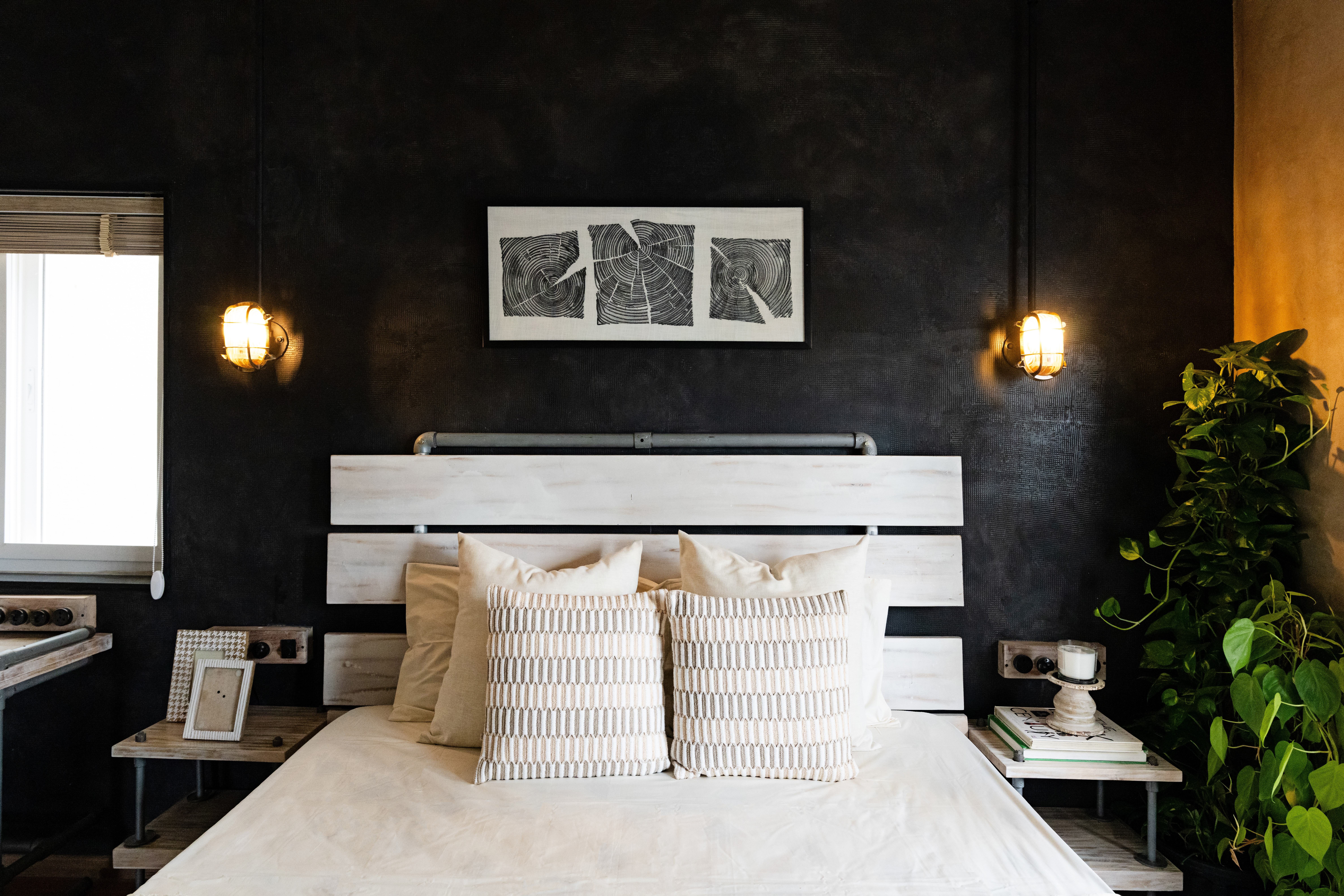 Bedroom with a black textured wall: whitewashed plank headboard on steel pipes, pale bedding with a symmetrical pair of patterned cushions, a framed woodcut triptych of tree rings above, two caged brass wall lamps, and trailing pothos to the right.